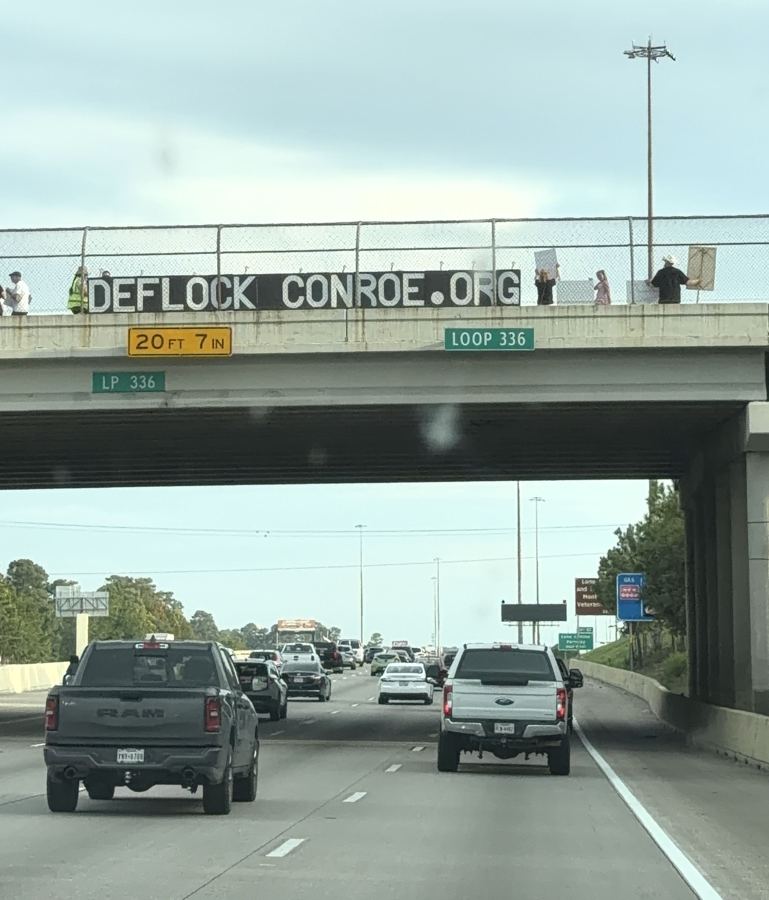 DEFLOCKCONROE.ORG banner hung over the N Loop 336 bridge, seen from the highway below